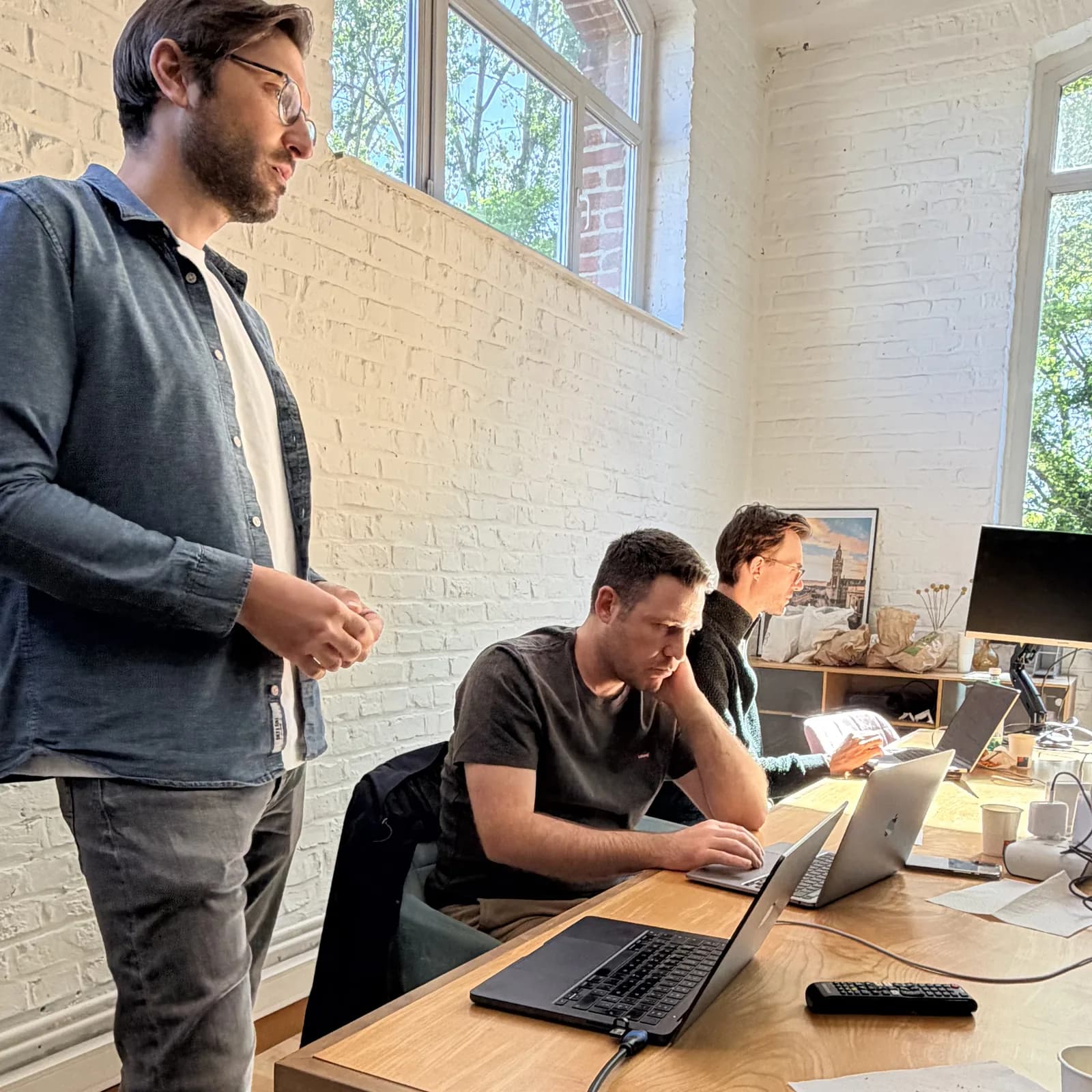 Atelier Hoko : un formateur debout accompagne deux développeurs concentrés sur leurs ordinateurs portables, dans un bureau aux murs de briques blanches.